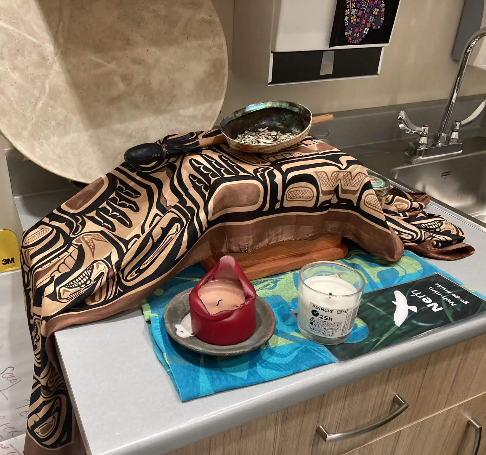 Traditional Indigenous items arranged on a countertop, including a large drum, a carved wooden piece covered with a patterned cloth, a bowl with sage, and two candles, in a healthcare setting with a sink and cabinets in the background.