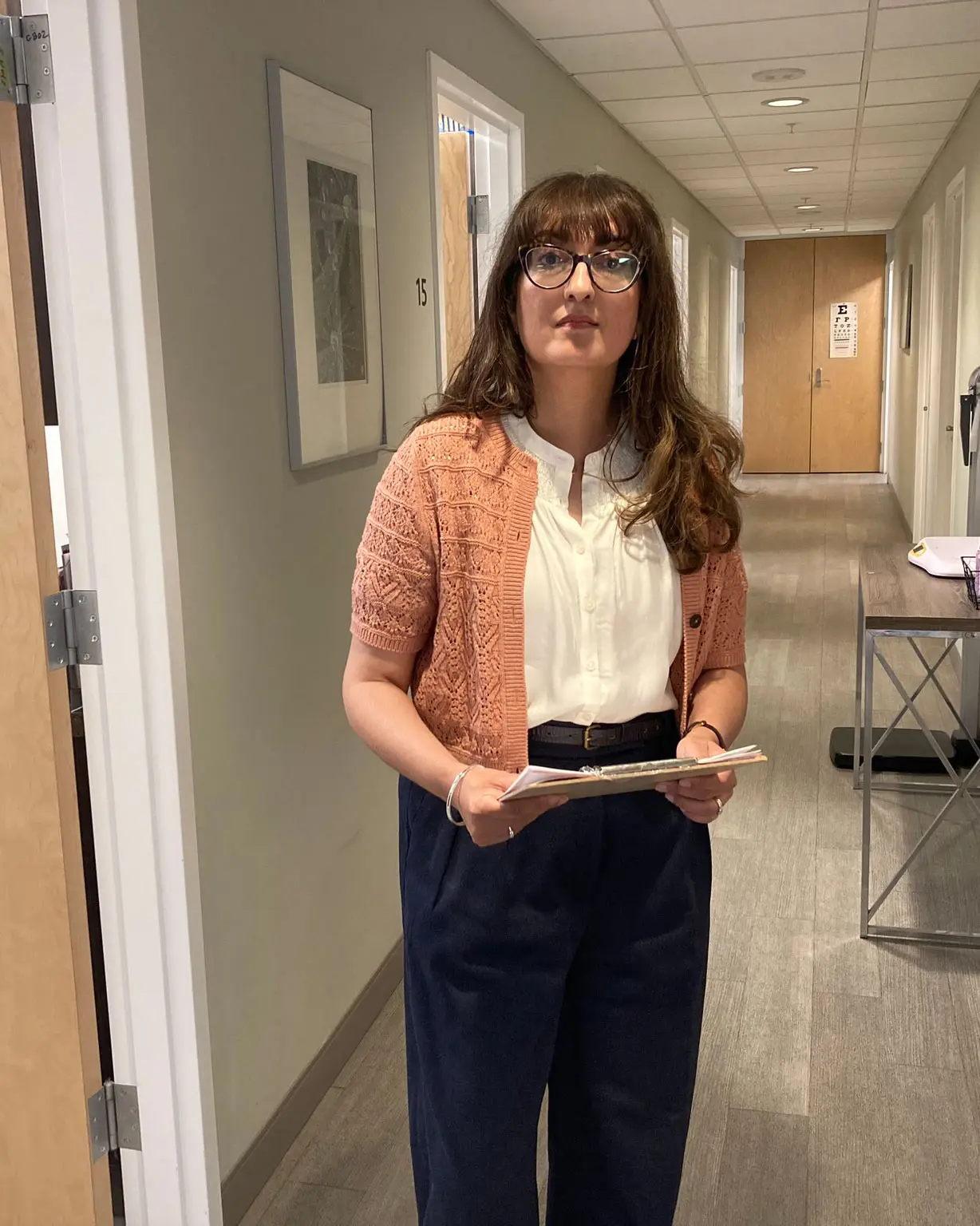 Dr. Micky Cooner standing in a hallway of a medical clinic, holding patient files, wearing glasses, a white blouse, and a peach cardigan, in South Surrey, BC.