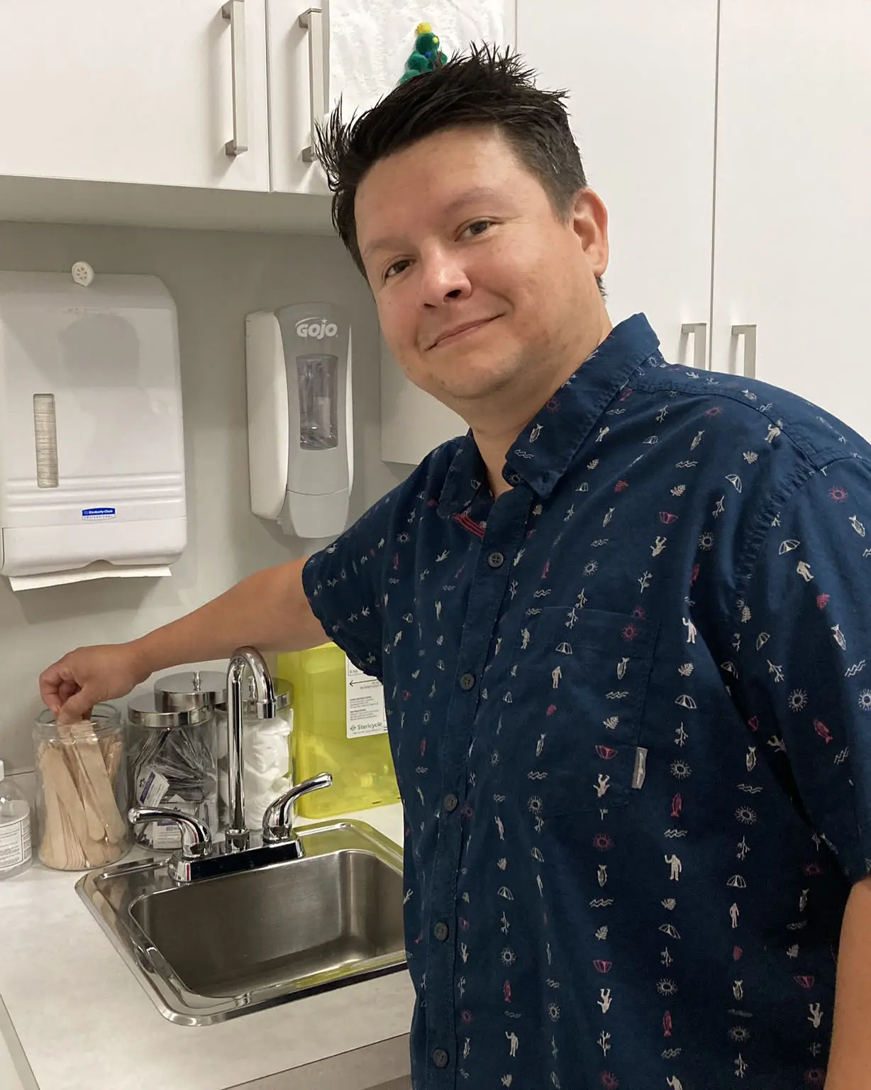 Dr. Toma Timothy standing next to a sink in a medical clinic, wearing a navy blue shirt with patterns, in White Rock, BC.