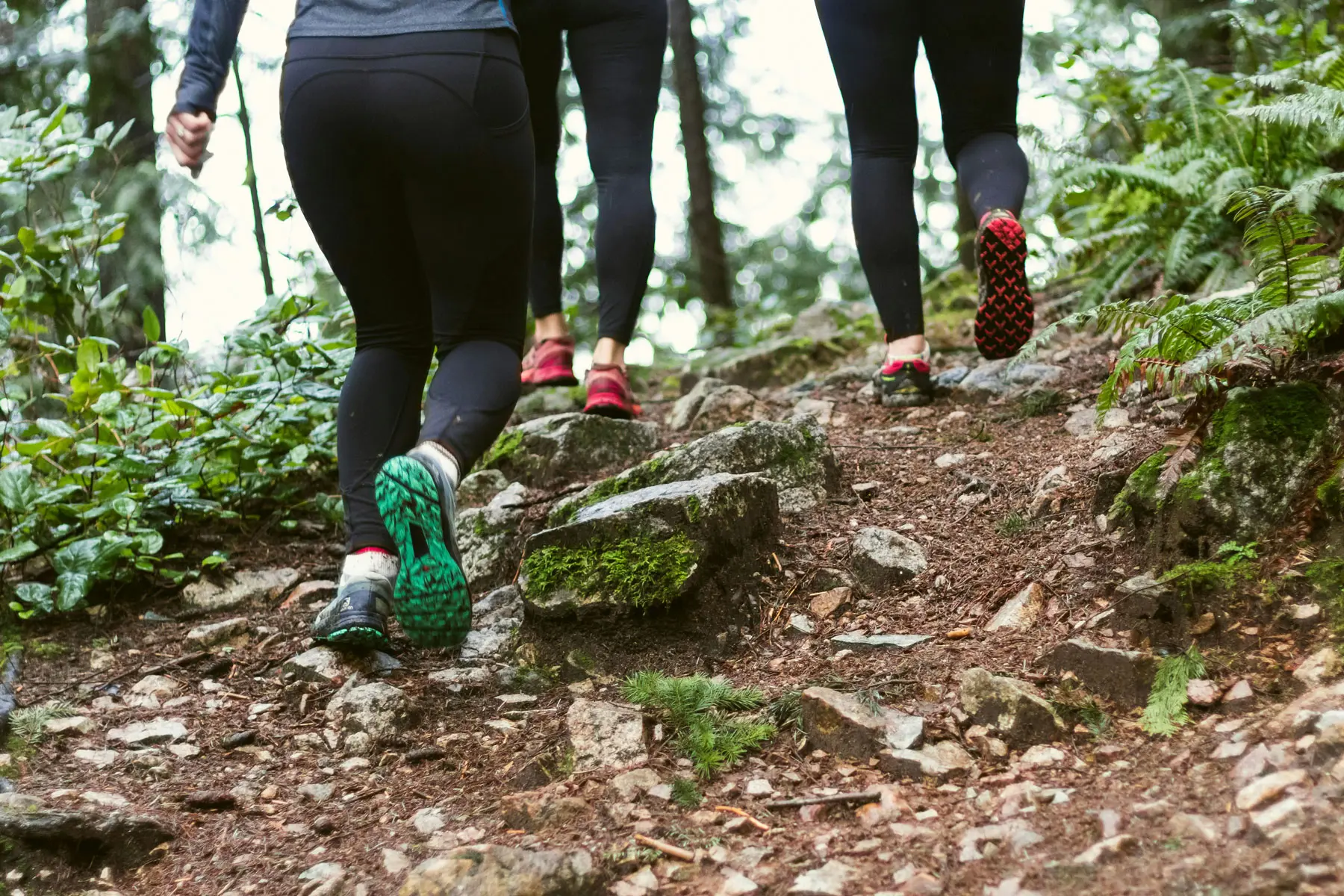 Close-up view of three people hiking up a rocky trail in a forest, showcasing their colorful running shoes and active lifestyle in White Rock South Surrey.