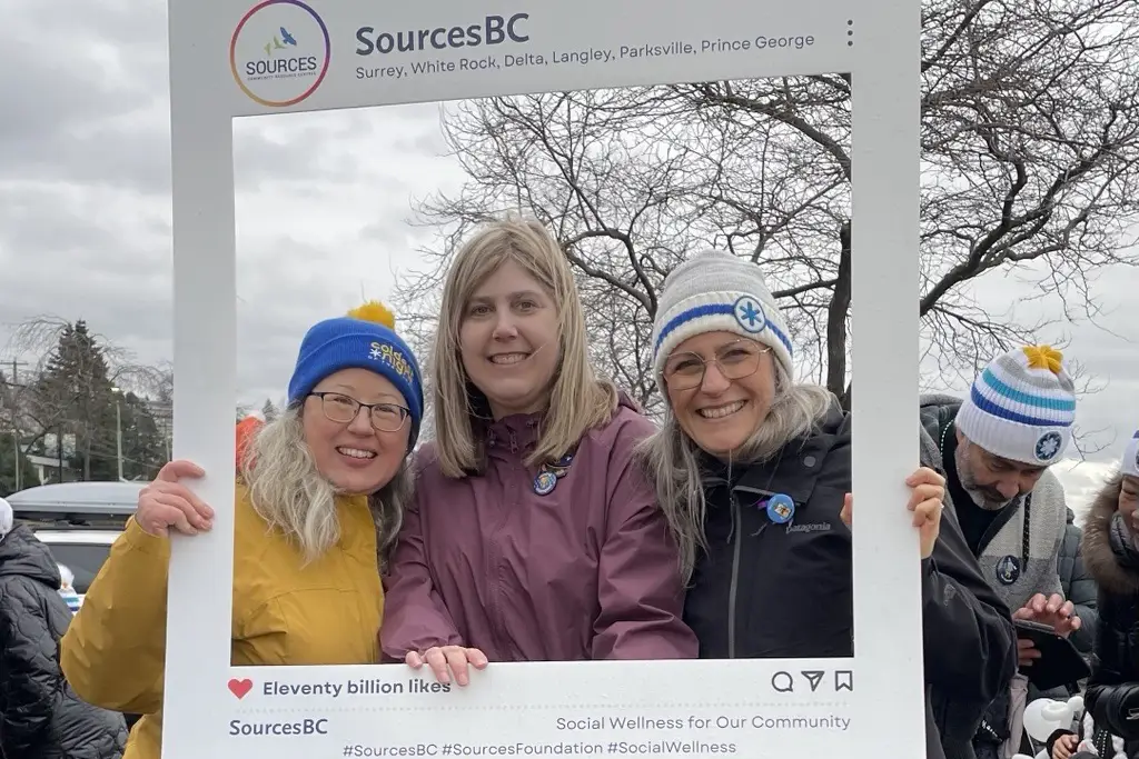 Three participants at a community event holding a SourcesBC frame, smiling and wearing winter hats and jackets, representing social wellness in Surrey, White Rock, Delta, 