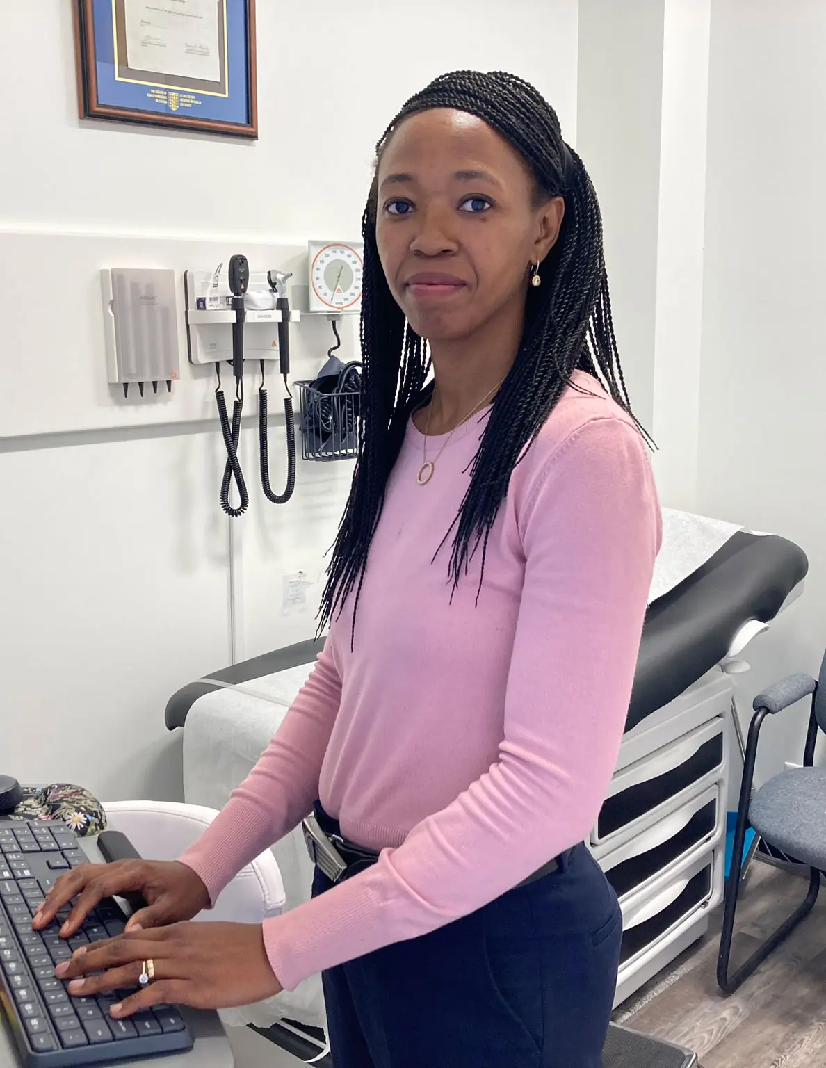 Dr. Abimbola Adewumi standing at a computer workstation in an examination room at Highroads Medical Clinic in South Surrey, BC, wearing a pink sweater.