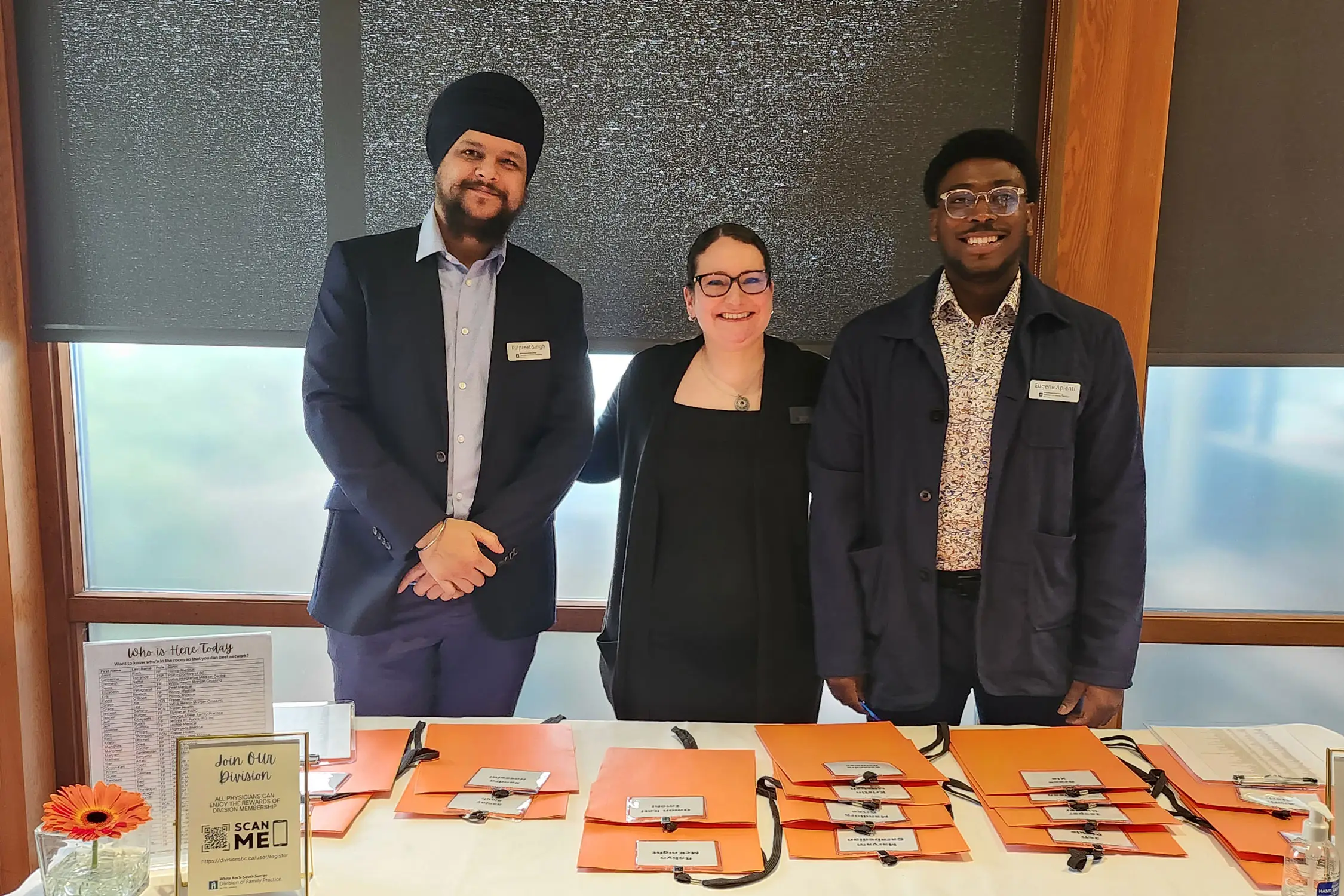 Three team members standing behind a registration desk with orange folders and name tags at a healthcare event in South Surrey White Rock